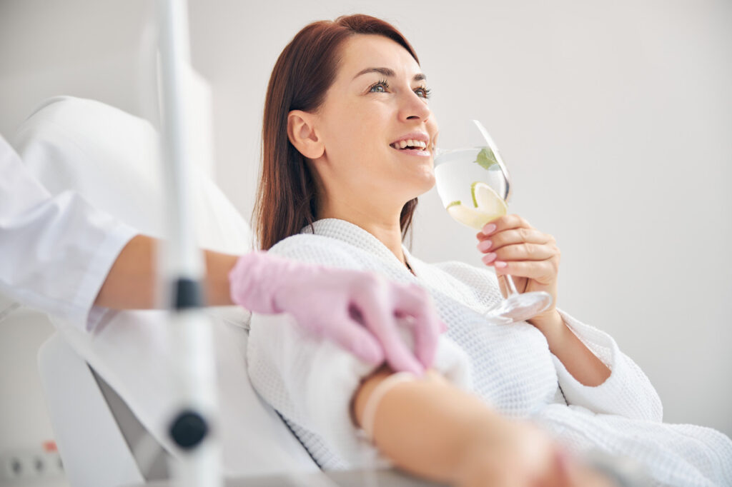 A woman smiles and drinks a glass of water while having a vitamin IV therapy treatment