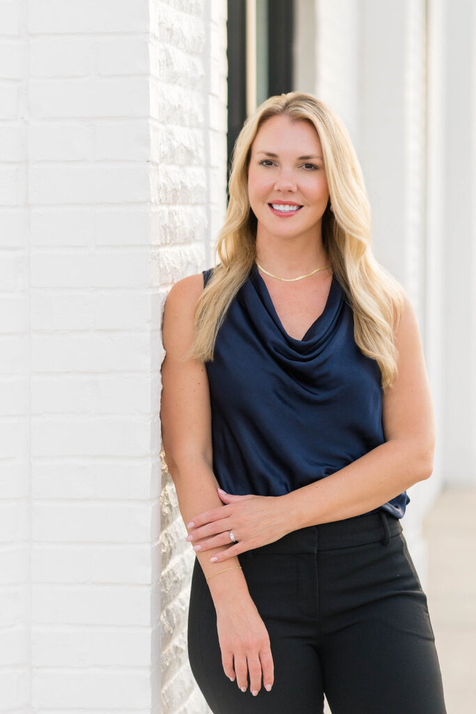 A blonde woman wearing an elegant navy blouse leans against a wall and smiles at the viewer